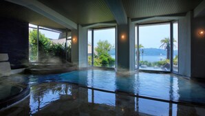 A modern indoor pool with large glass doors offering views of a tropical landscape and a body of water. - Kurofune Hotel (Shimoda)