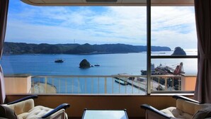 A room with a large window offering a view of a calm sea, boats, and distant land. There are two armchairs and a small table in the foreground. - Kurofune Hotel (Shimoda)