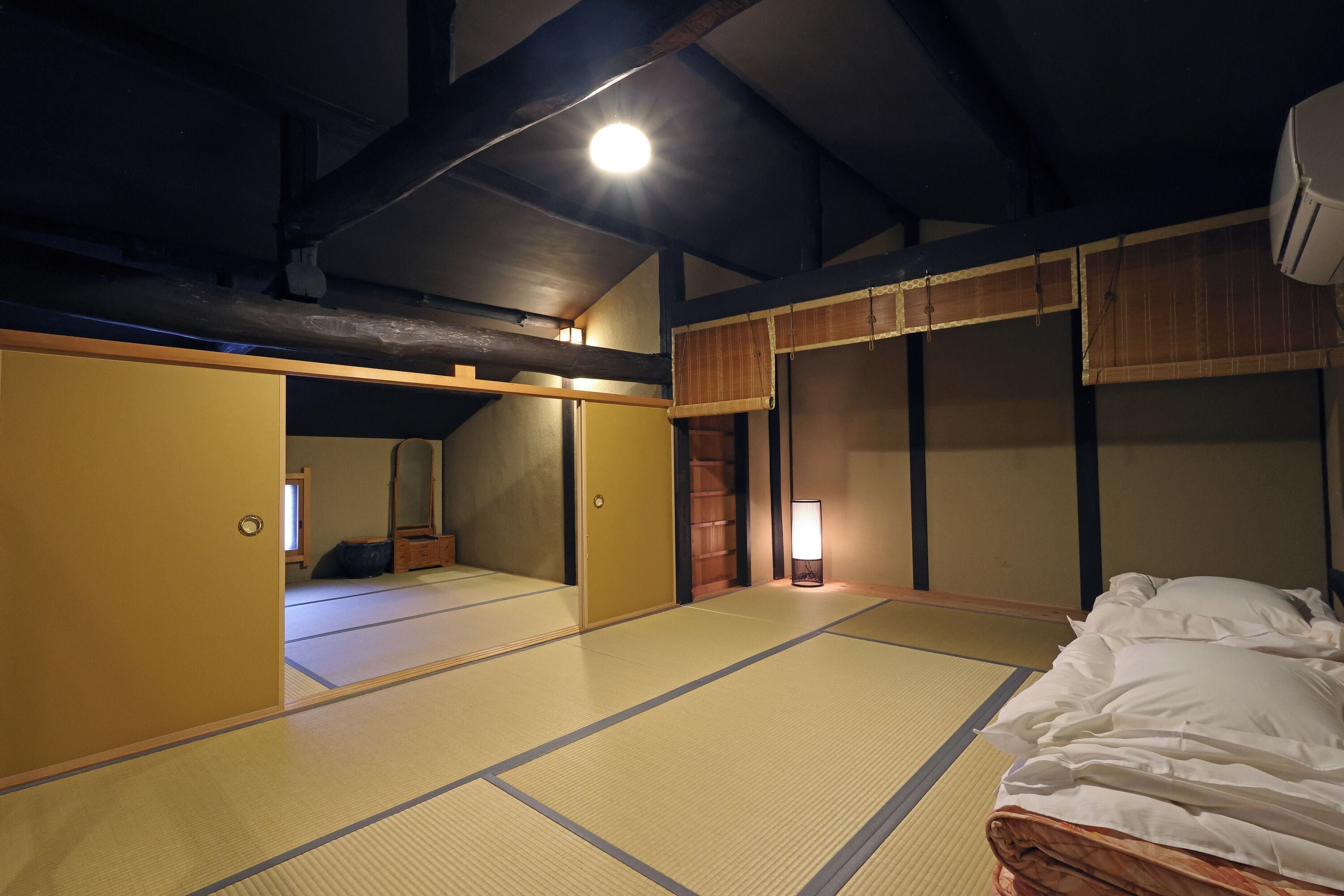 A traditional Japanese room with tatami flooring, a bed with white bedding, a nightstand, and a wall-mounted air conditioner.