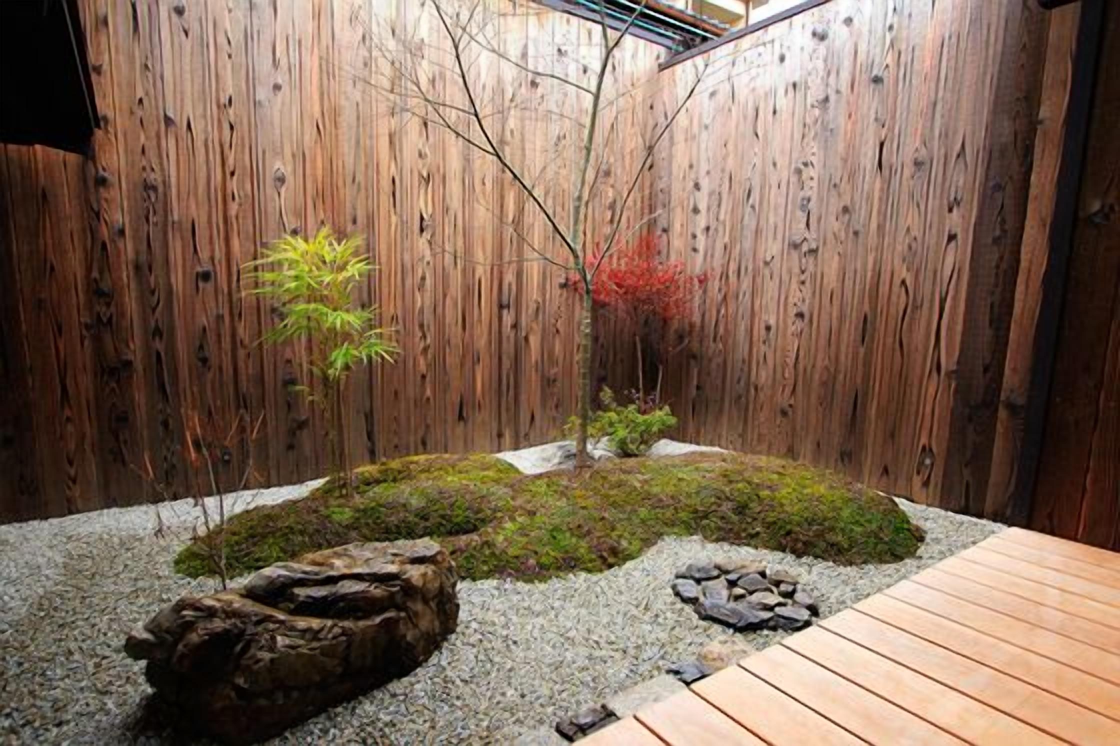 Una valla de madera, un árbol pequeño, musgo, rocas y una mesa de madera.