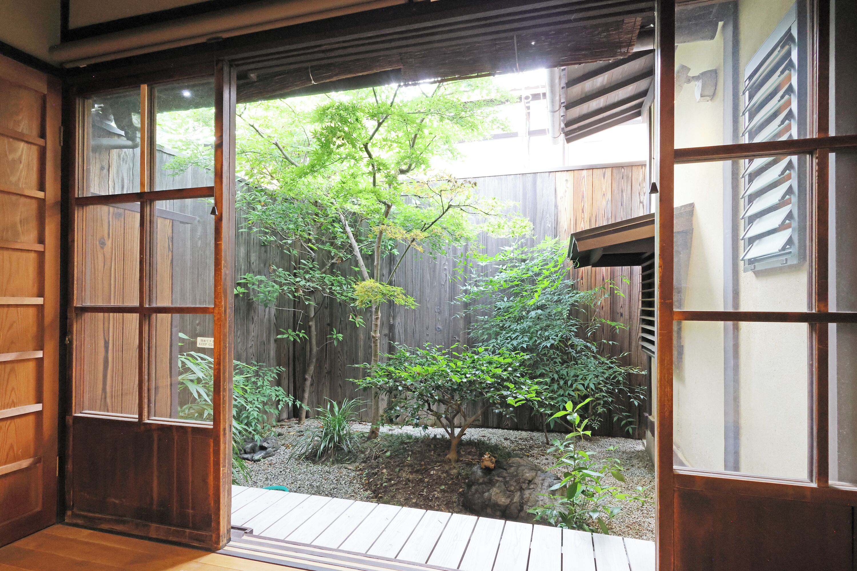 Une pièce de style japonais traditionnel, avec un sol recouvert de tatamis, des portes coulissantes en papier de riz (shoji) et une vue sur un jardin agrémenté d’un petit arbre et d’un chemin de gravier.