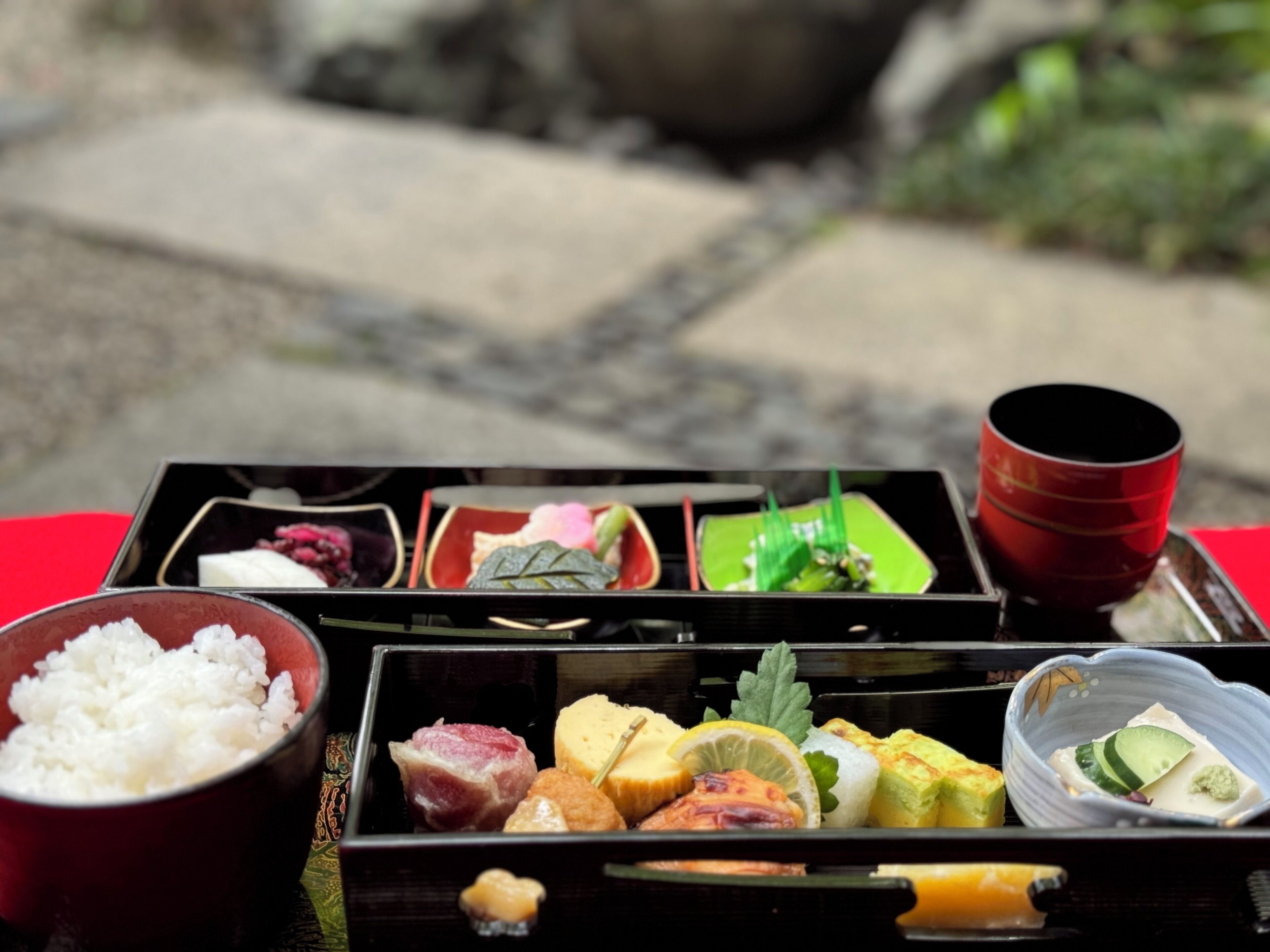 A traditional Japanese meal setup with various dishes in separate compartments, a bowl of rice, and a red cup.