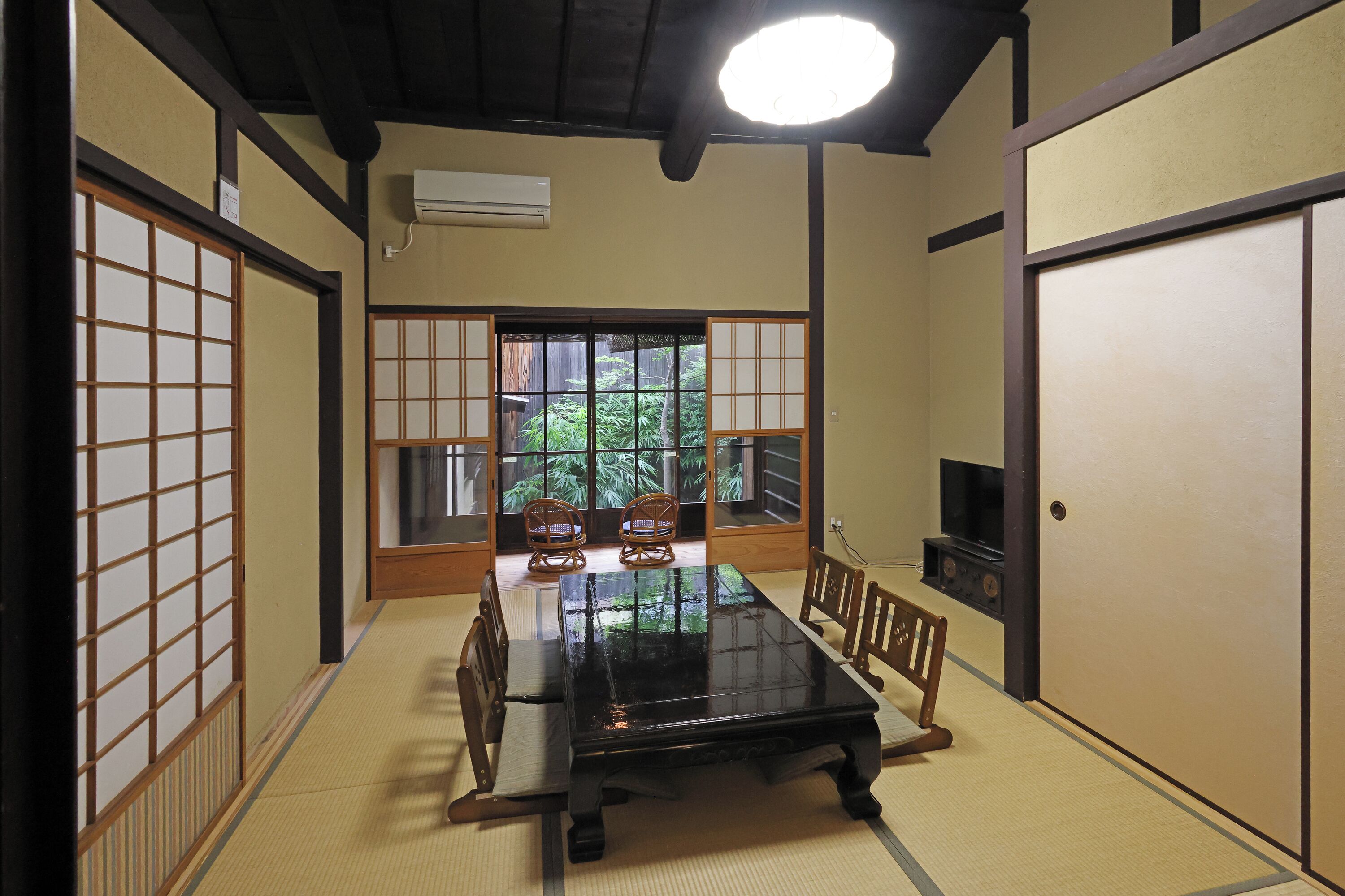 A traditional Japanese room with tatami flooring, a black lacquered table, wooden chairs, sliding doors, and a TV.