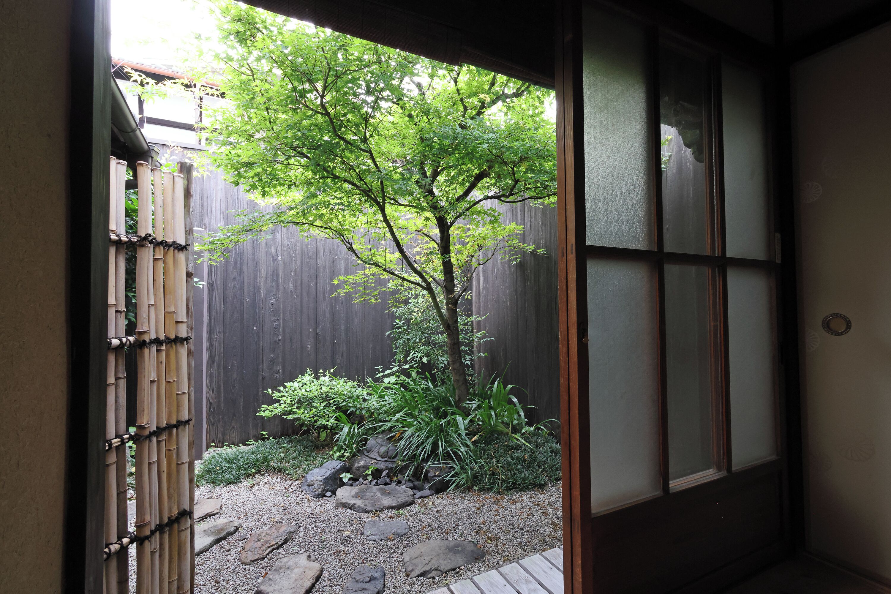 Une pièce de style japonais traditionnel, avec un sol recouvert de tatamis, une porte coulissante en papier de riz (shoji) et une vue sur un jardin agrémenté d’une clôture en bambou et d’un arbre.