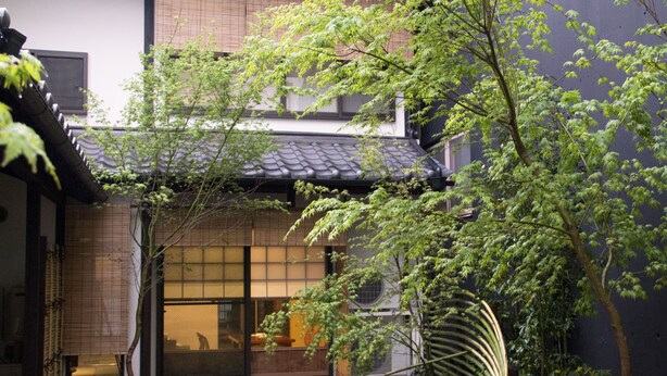 Un edificio japonés tradicional con tejado de tejas, entrada de madera y un pequeño jardín con un árbol y un farol de piedra.