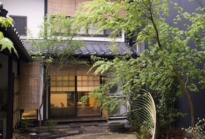 Ein traditionelles japanisches Gebäude mit einem Ziegeldach, einem hölzernen Eingang und einem kleinen Garten mit einem Baum und einem Steinglaternen.