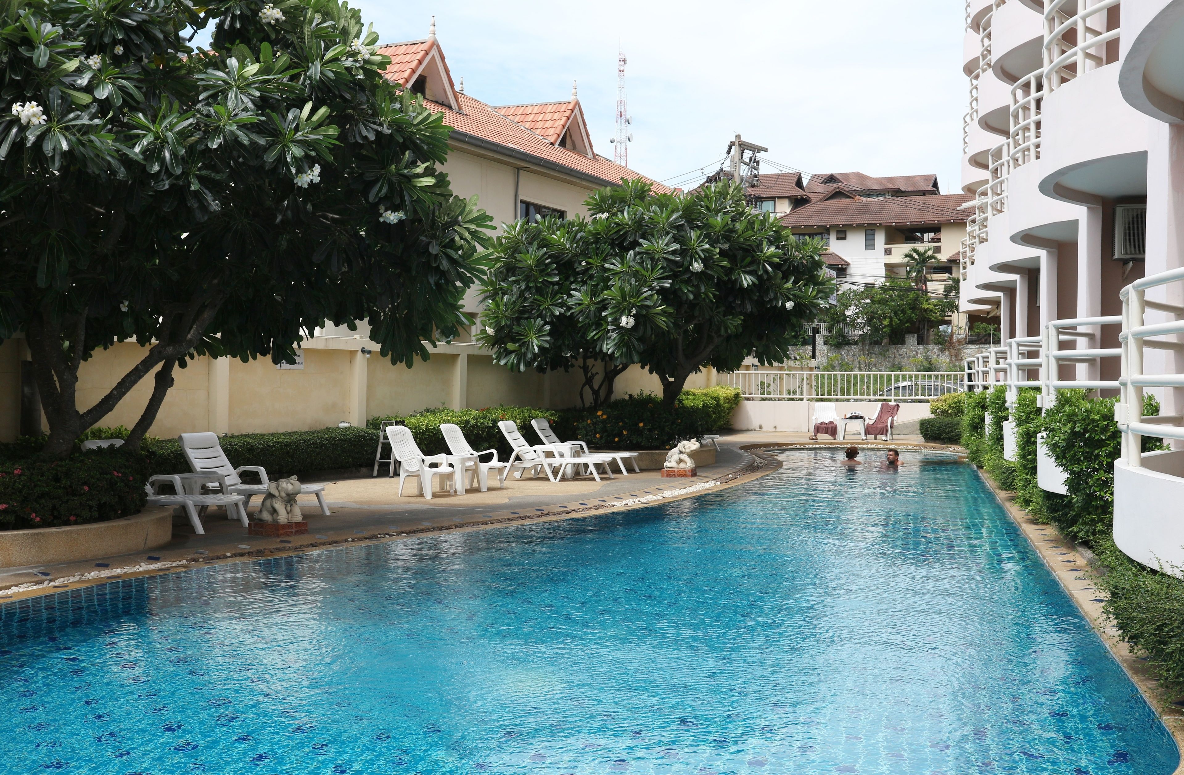 A swimming pool with lounge chairs, surrounded by greenery and buildings.