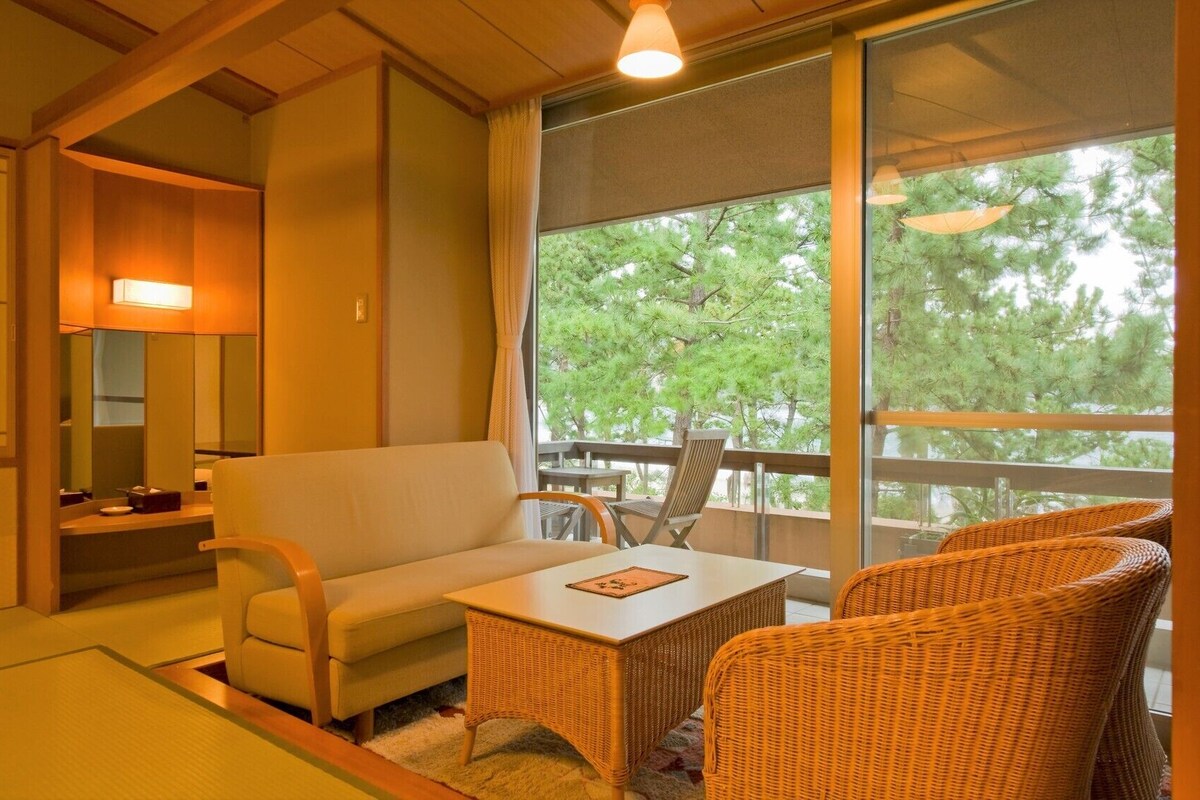 A room with a sofa, a table, wicker chairs, a balcony with a view of trees, and a large window.