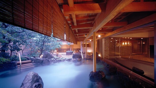 A traditional Japanese onsen with a stone bath surrounded by rocks, wooden beams, and a view of a traditional building. - Ryokan Tokiichiyu (Minamiizu)
