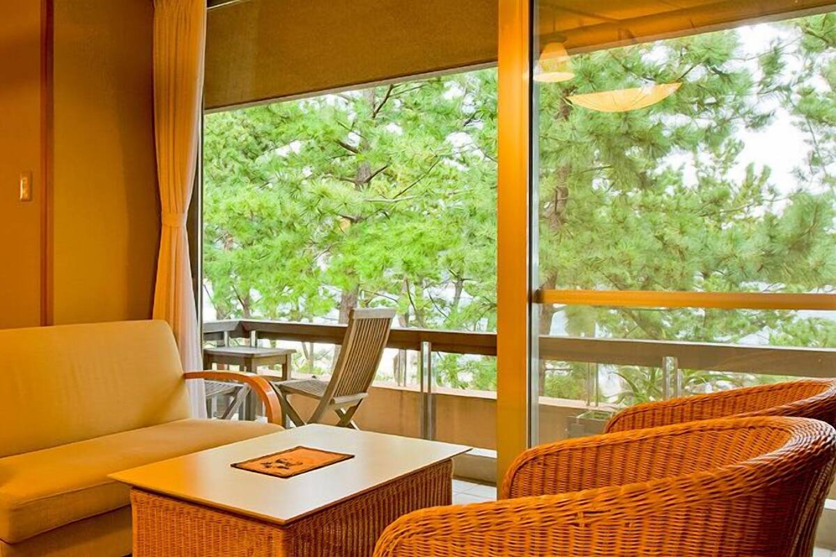 Quarto com sofá, mesa pequena, cadeiras de vime, varanda com vista para árvores e espelho.