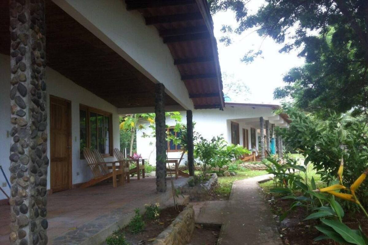 Un espacio al aire libre techado con pilares de piedra, mobiliario de madera y un camino que lleva a un edificio.