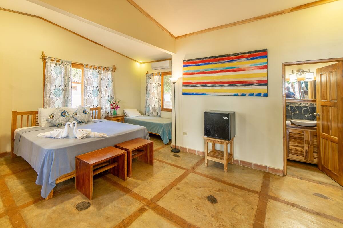 Un dormitorio con cama, un banco, televisión y un baño visible a través de una puerta abierta.