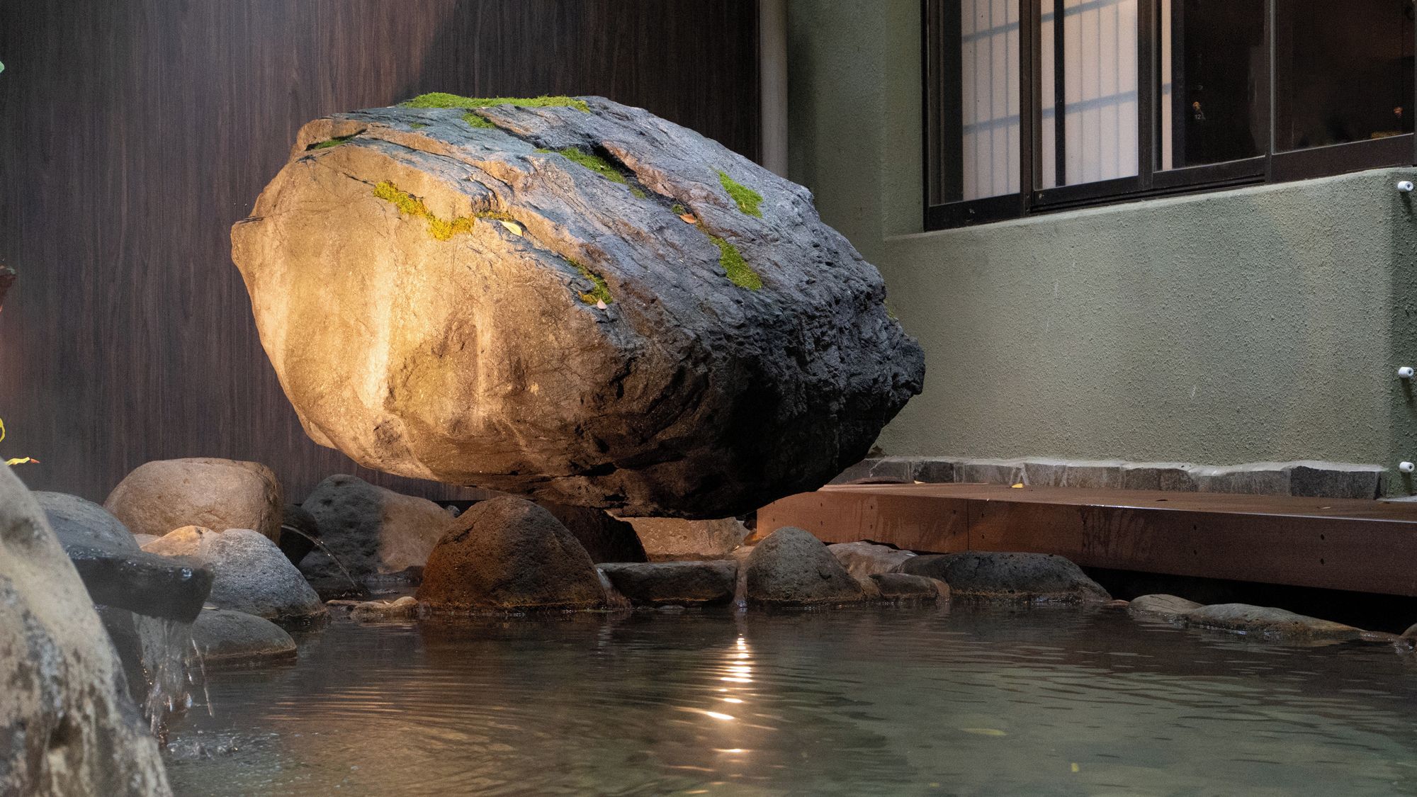 A rock balanced on a smaller rock over water, with a building in the background.