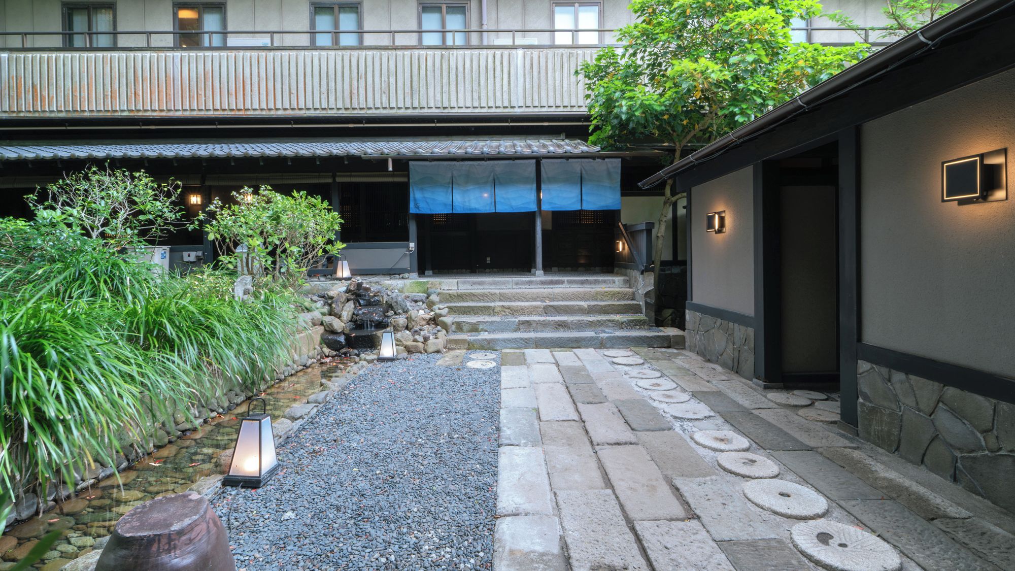 A courtyard with a stone pathway leading to a building entrance, surrounded by greenery and a small water feature.