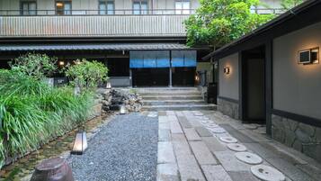 A courtyard with a stone pathway leading to a building entrance, surrounded by greenery and a small water feature.