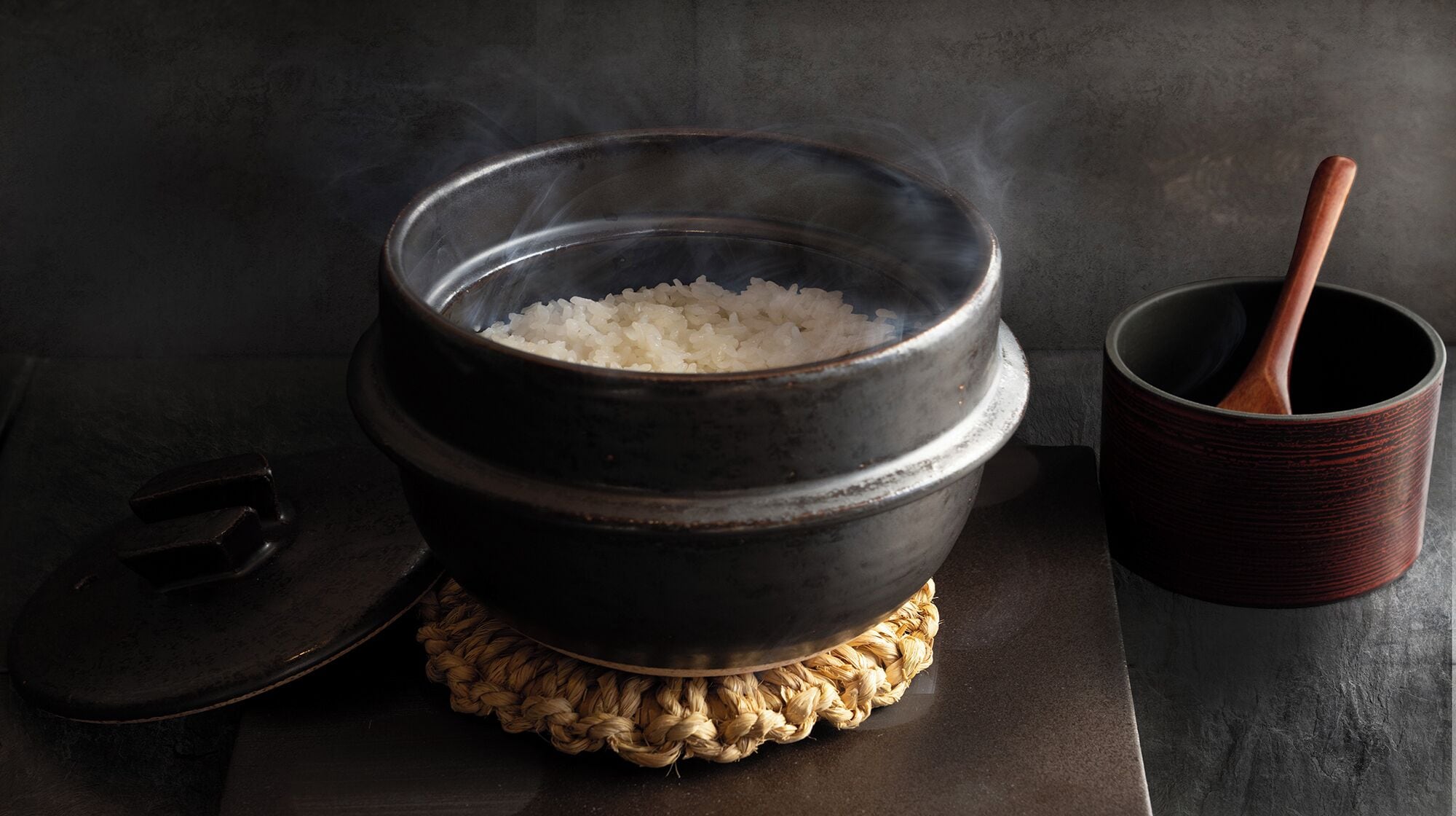 A pot of rice on a woven mat, a wooden spoon, and a dark textured surface.