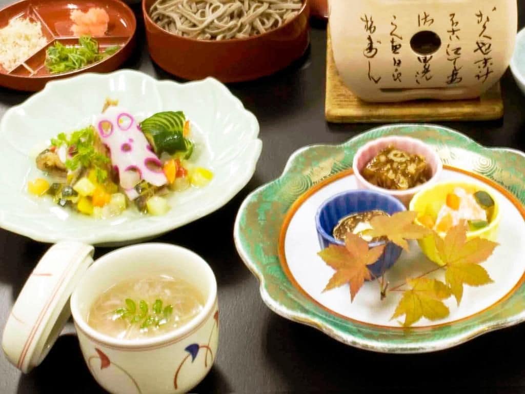 Una mesa con varios platos, incluyendo un tazón de fideos, un plato de verduras y una taza de sopa.