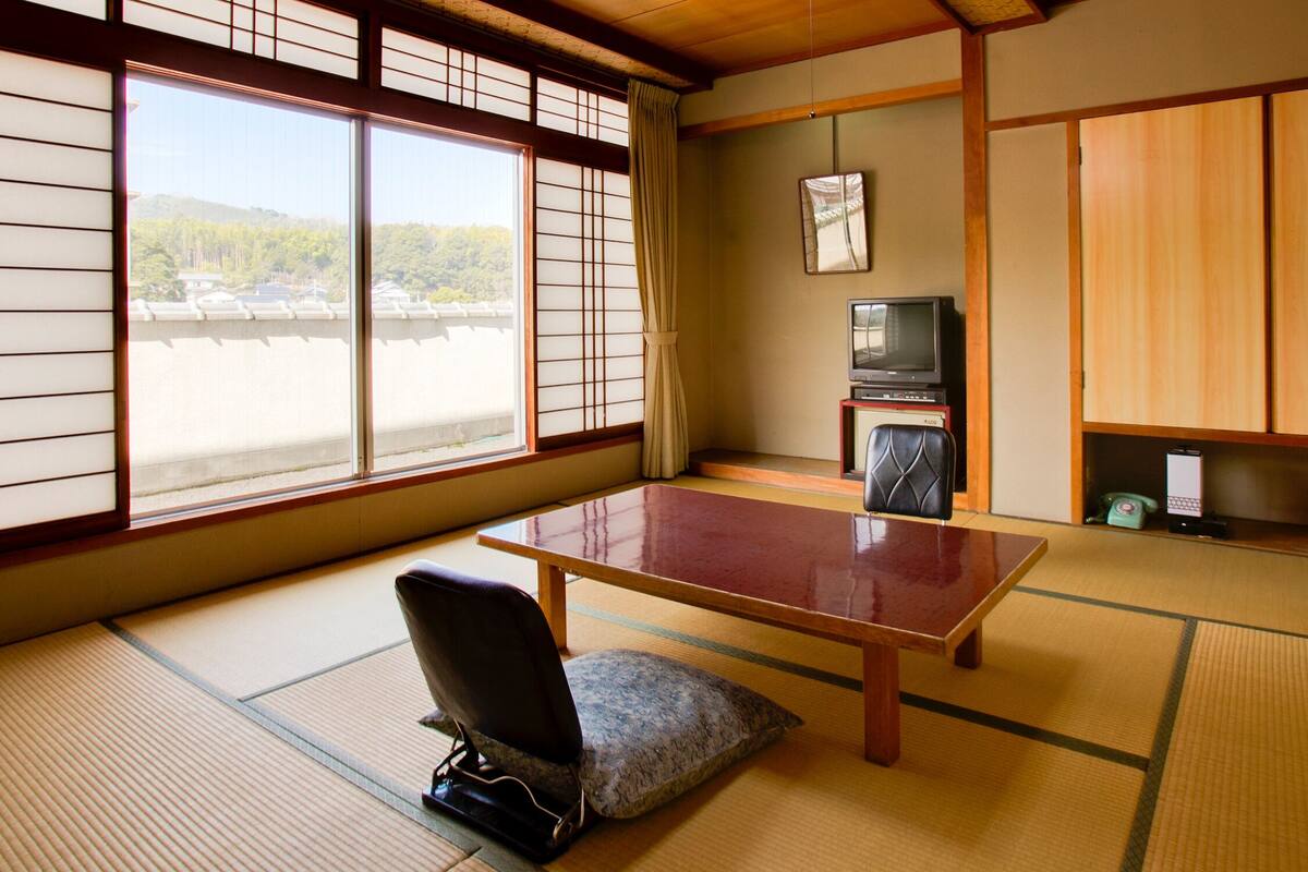 Une pièce de style japonais traditionnel, avec un sol recouvert de tatamis, une table basse, une chaise, un téléviseur et des portes coulissantes.