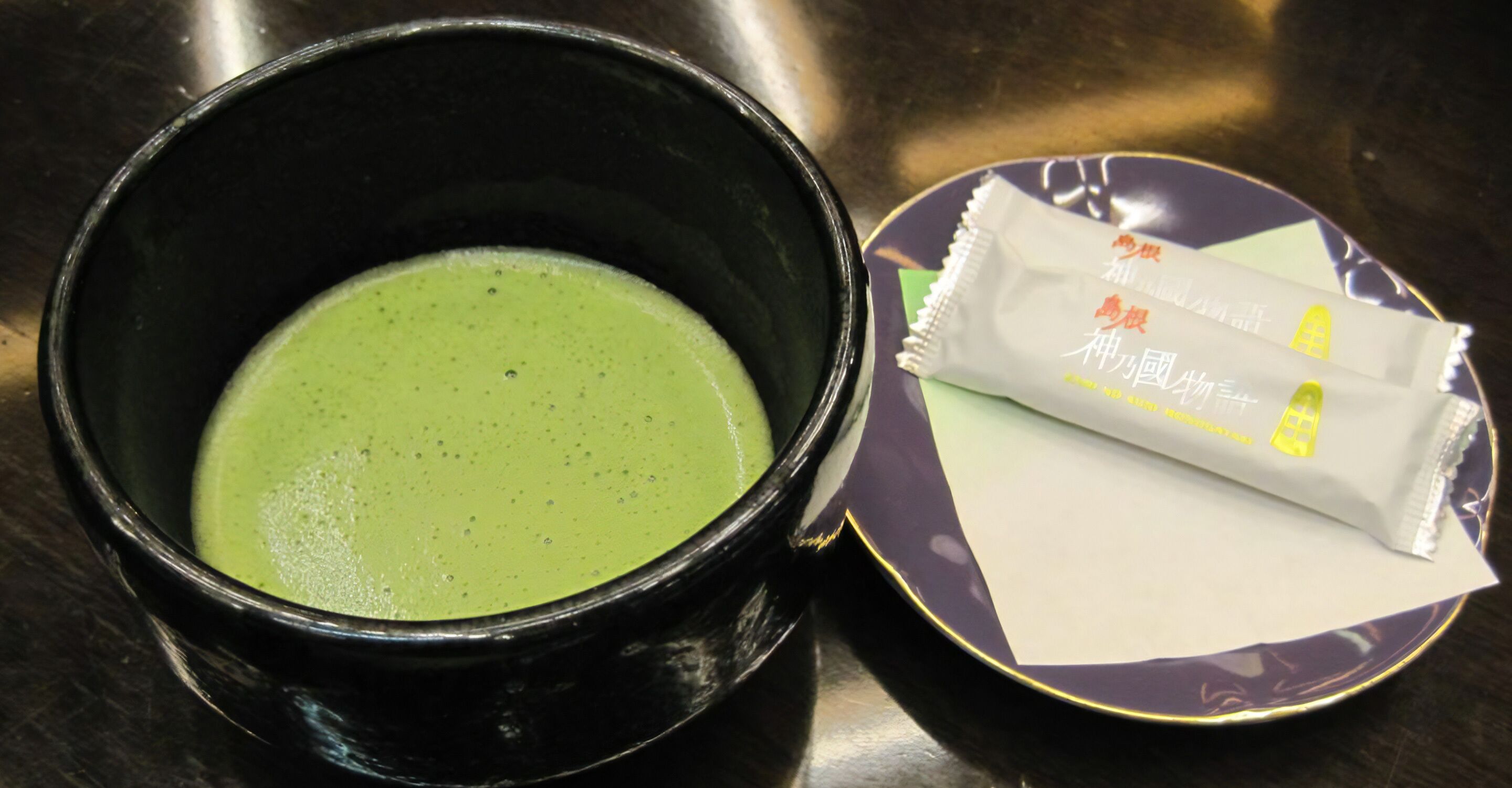A black bowl with a green liquid, a purple plate with a white packet, and a spoon.