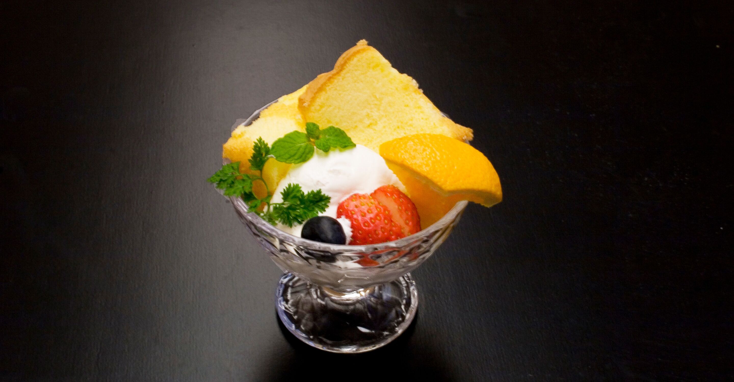 A dessert in a clear glass bowl with a slice of cake, strawberries, blueberries, a lemon slice, and a dollop of cream.