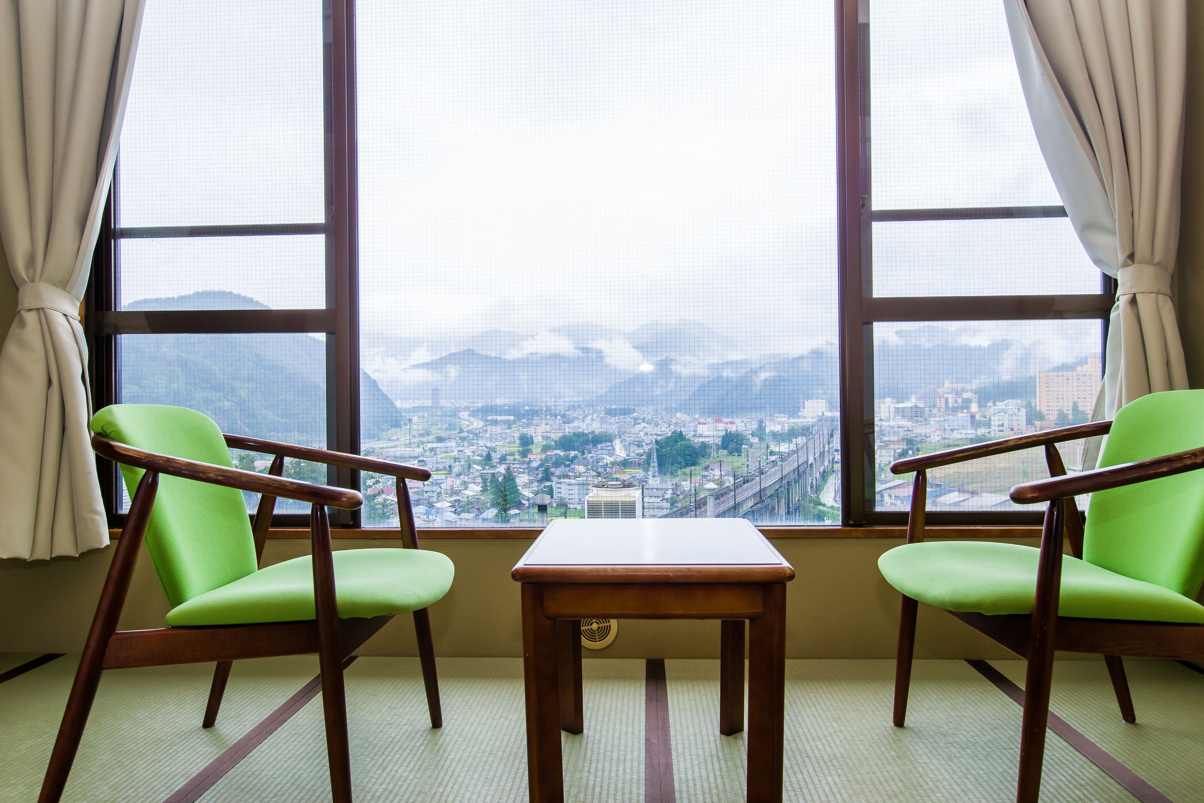 A room with two green chairs and a small wooden table, large windows with a view of a city and mountains.