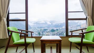 A room with two green chairs and a small wooden table, large windows with a view of a city and mountains.
