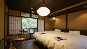 A traditional Japanese-style room with tatami flooring, a low wooden table, a single wooden chair, a large bed with white bedding, and a view through a shoji screen to the outside. - Rinn Shijo Takasegawa (Kyoto)
