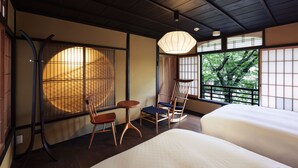 Perinteinen japanilaistyylinen huone, jossa on tatamimatto, matala puupöytä, pehmustettu tuoli, sänky ja suuri ikkuna, josta avautuu näkymä puita kohti.