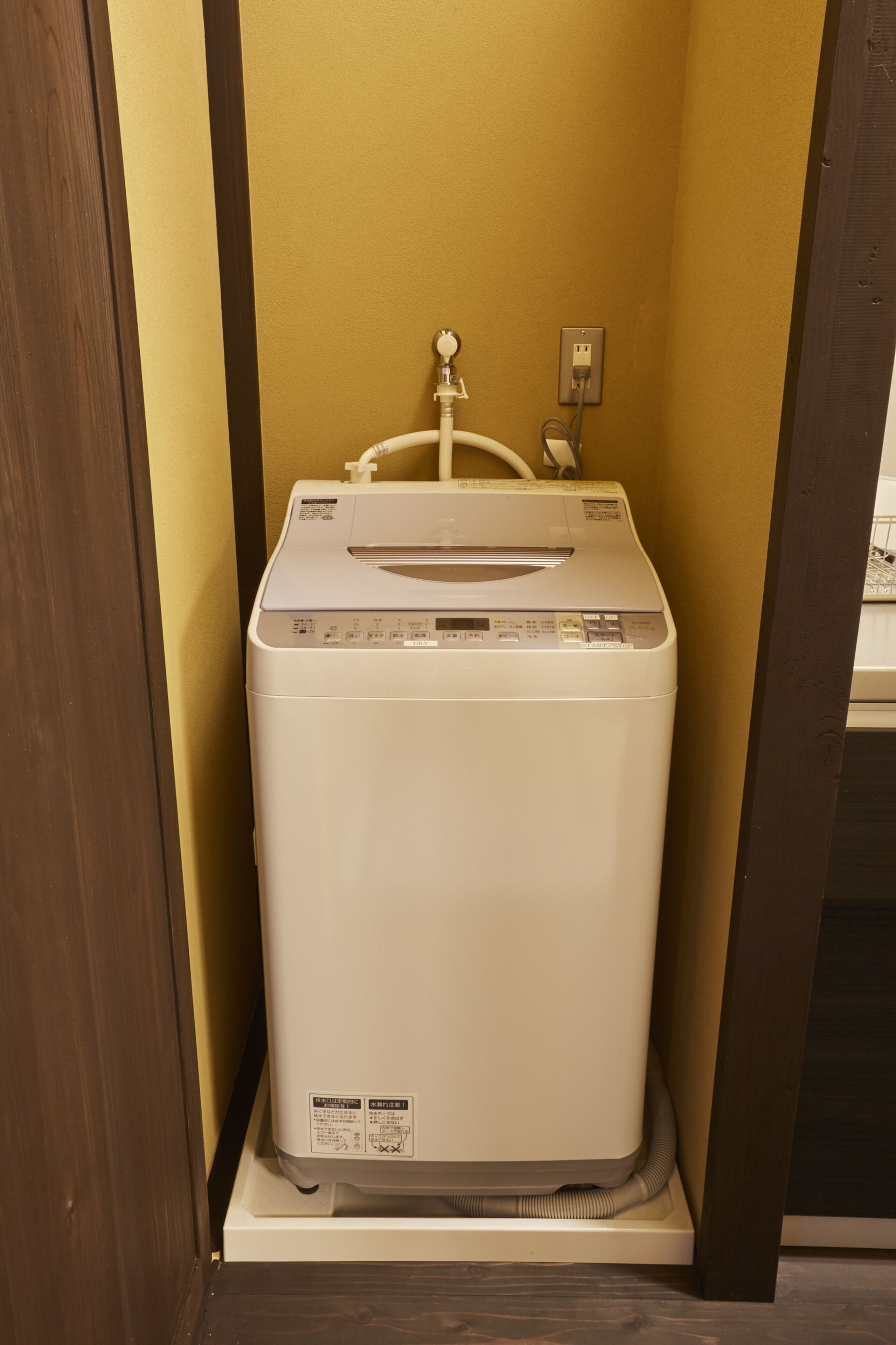 A compact, white, wall-mounted washer and dryer unit in a corner with a wooden door frame and a yellow wall.