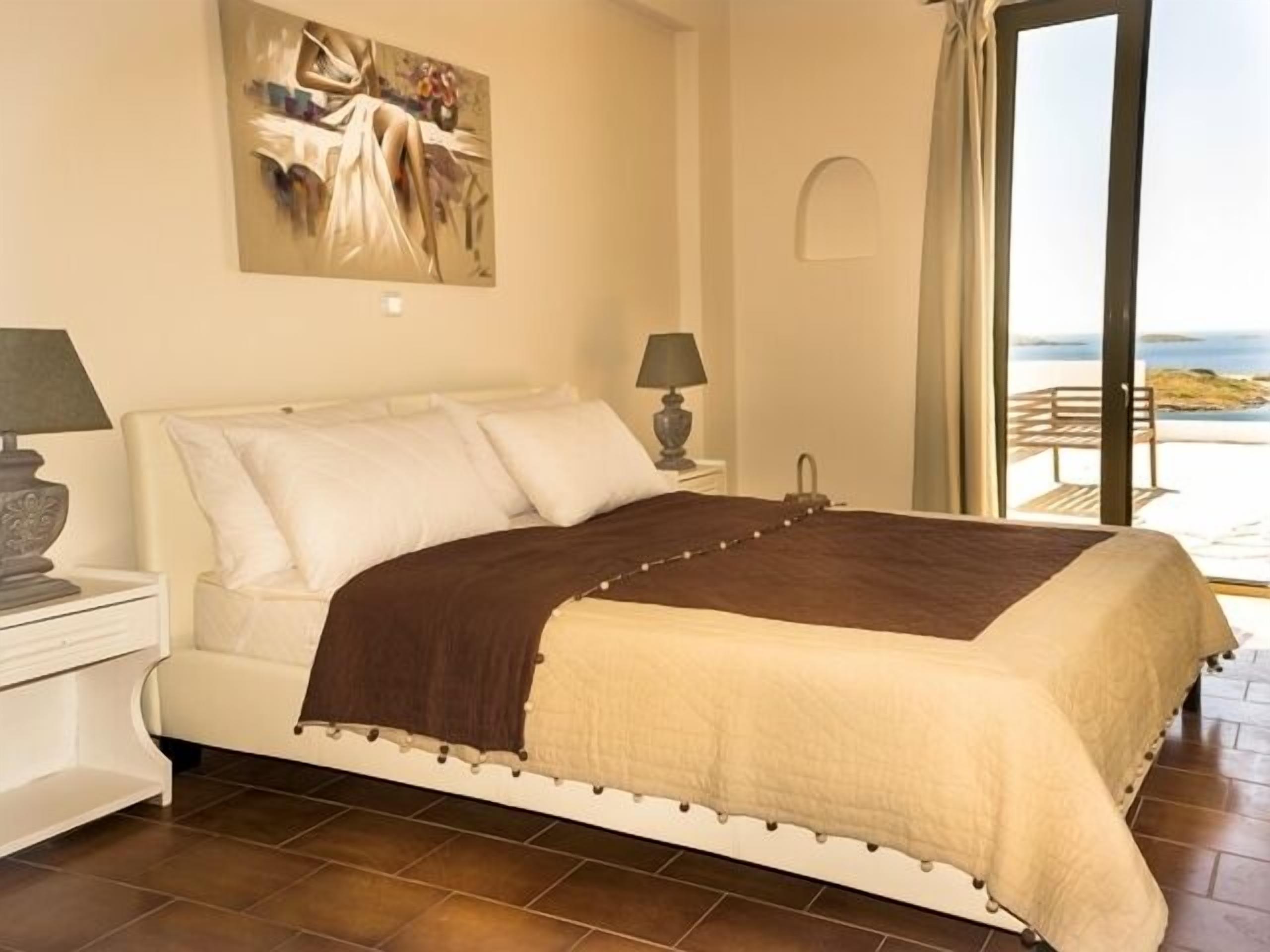 A bedroom with a bed, bedside tables, a painting, and a view of the sea.