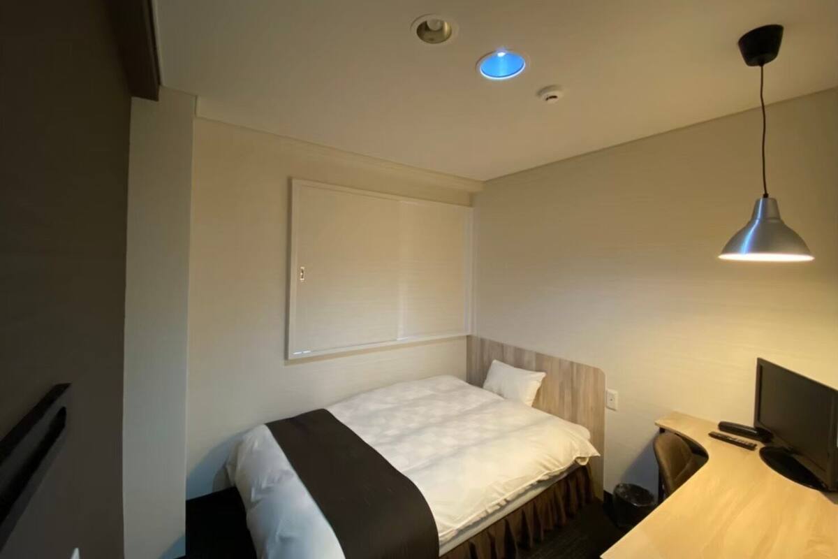 A hotel room with a bed, a desk, a chair, a TV, and a ceiling light.