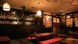 Een schemerig verlichte bar met houten meubels, flessen op planken en hangende lampen.
