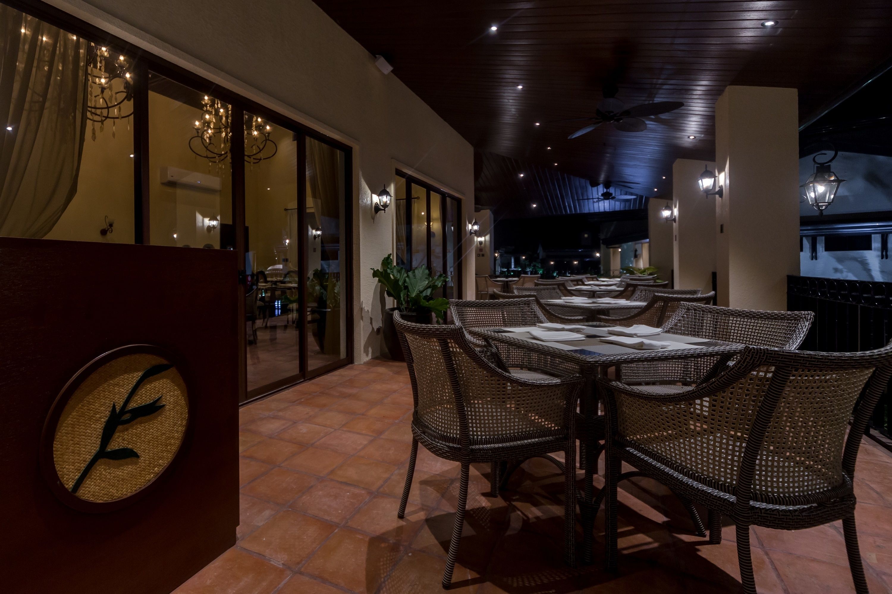 An outdoor dining area with wicker tables and chairs, a clock on the wall, and a view of the interior through glass doors.
