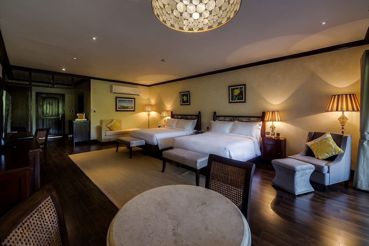 Una habitación de hotel con una cama grande, dos mesitas de noche, una mesa redonda, una silla y una lámpara decorativa en el techo.
