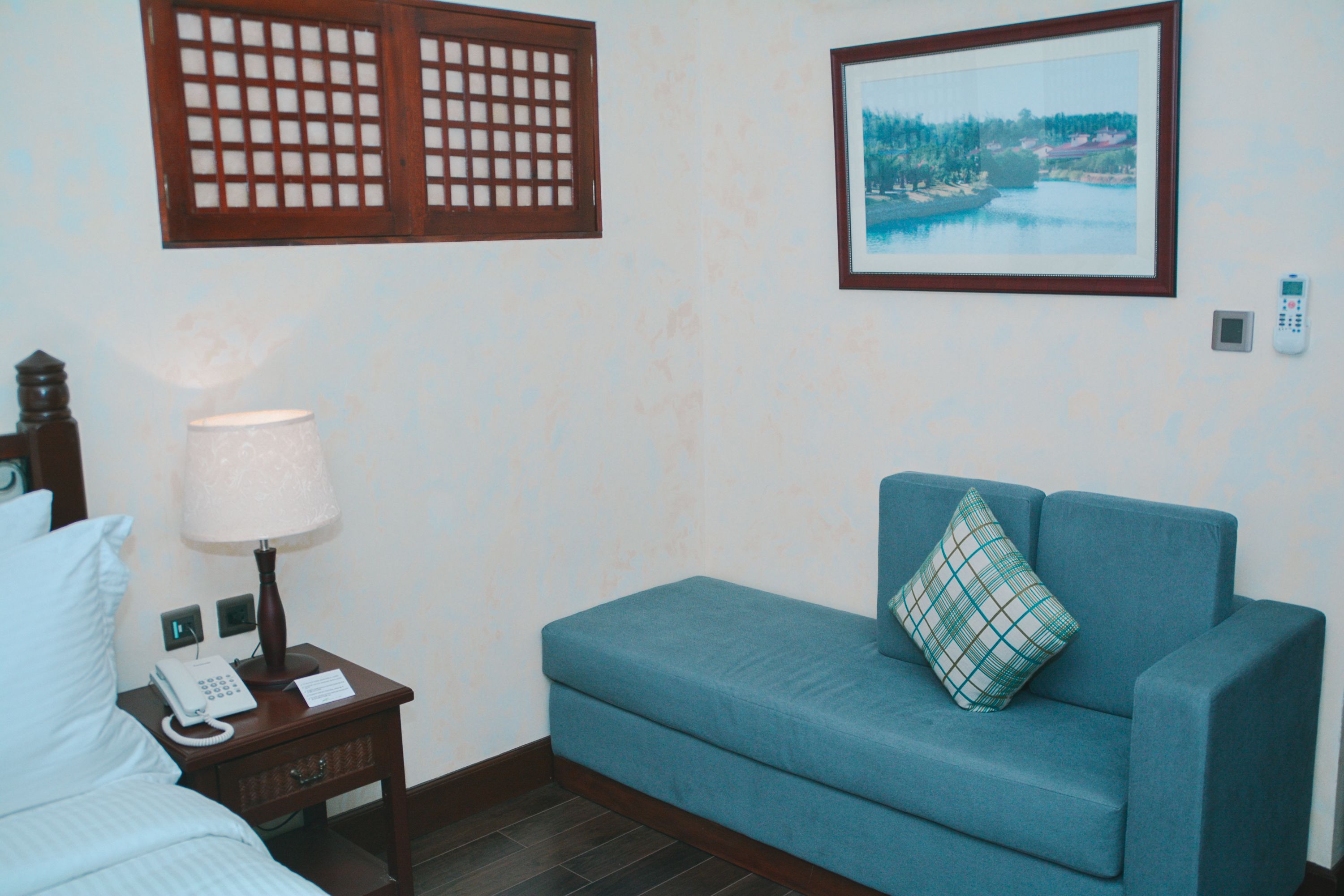 A hotel room with a bed, a nightstand with a lamp and phone, a blue sofa with a pillow, a picture on the wall, and a window with a view of trees and water.