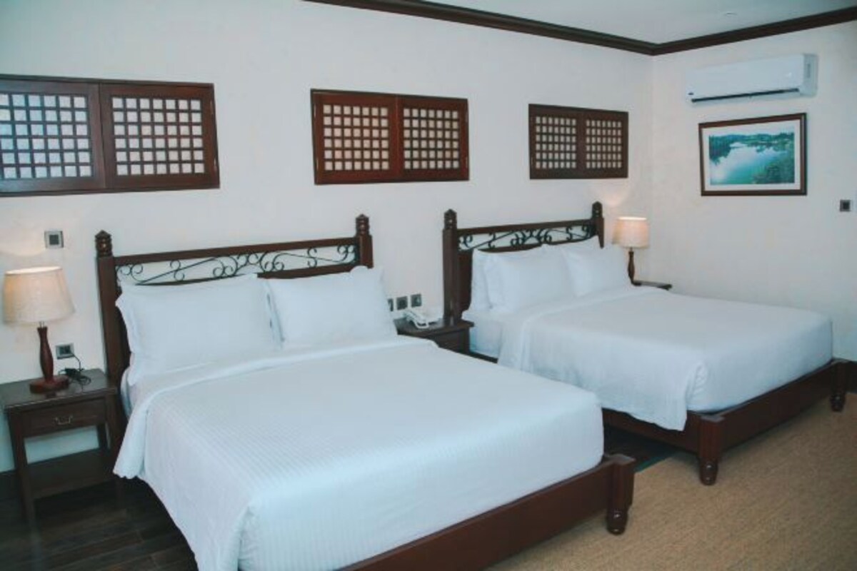 A hotel room with two beds, wooden headboards, a nightstand with a lamp, and a wall-mounted air conditioner.