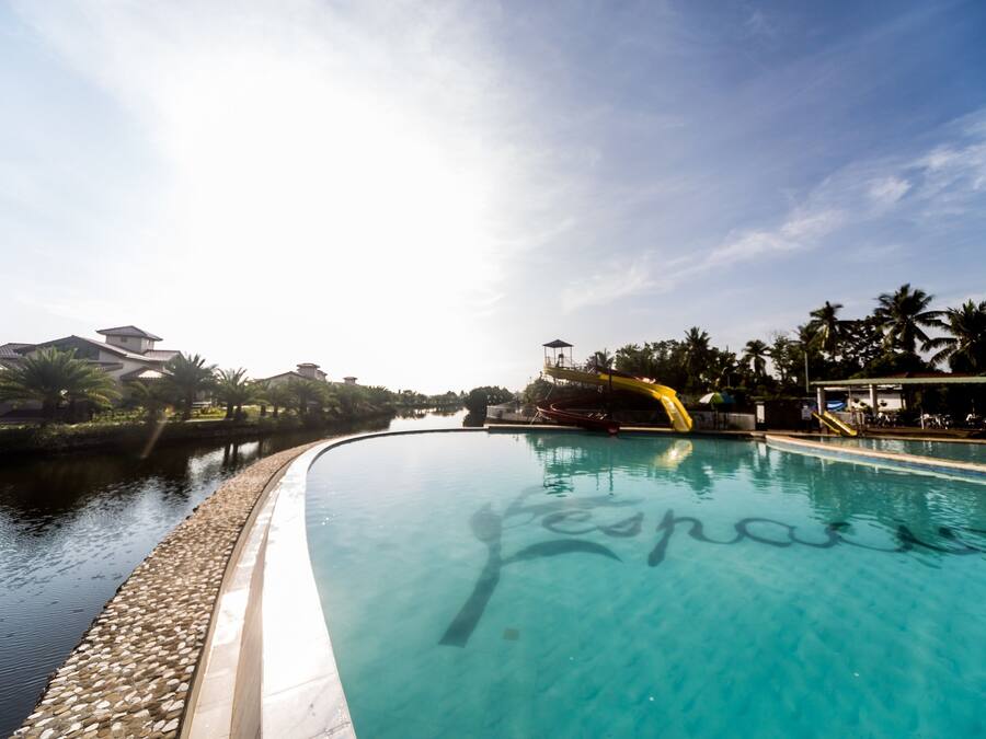Uma piscina transparente com um tobogã, rodeada por palmeiras e edifícios.