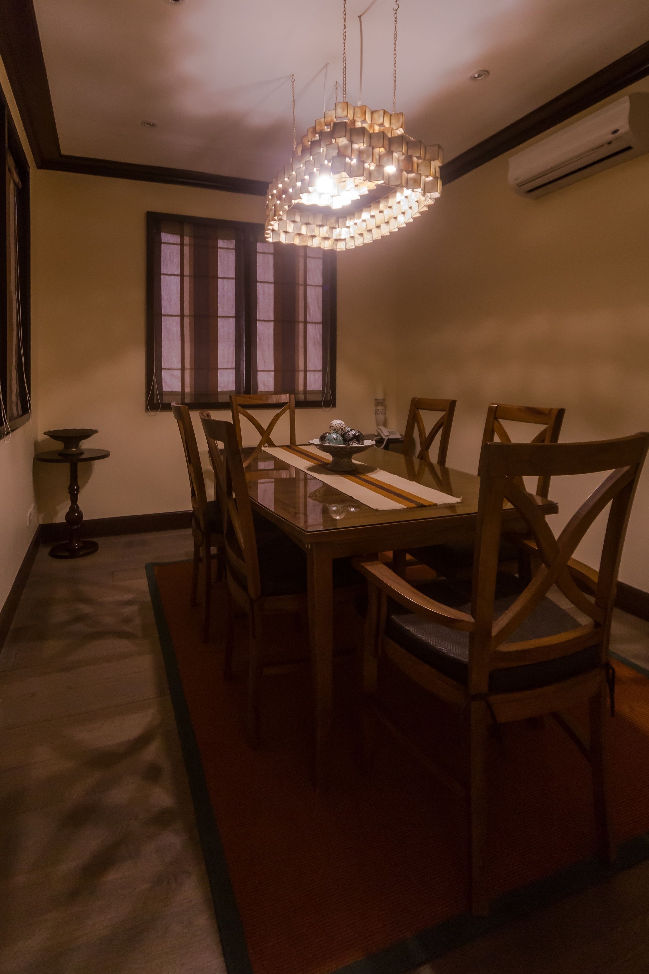 Un comedor con una mesa rectangular, seis sillas de madera, una lámpara colgante, una ventana con cortinas y un equipo de aire acondicionado en la pared.