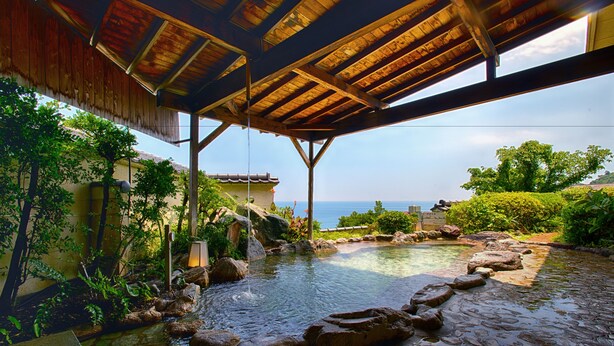 A stone water feature with a wooden deck overhead, surrounded by greenery and a view of the sea. - Atagawa Prince Hotel (Higashiizu)
