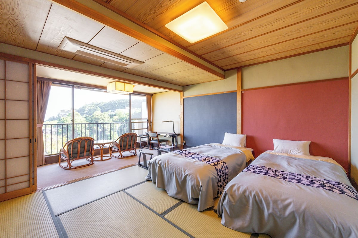 A traditional Japanese room with tatami flooring, two beds, a desk, and a balcony with a view of the mountains.