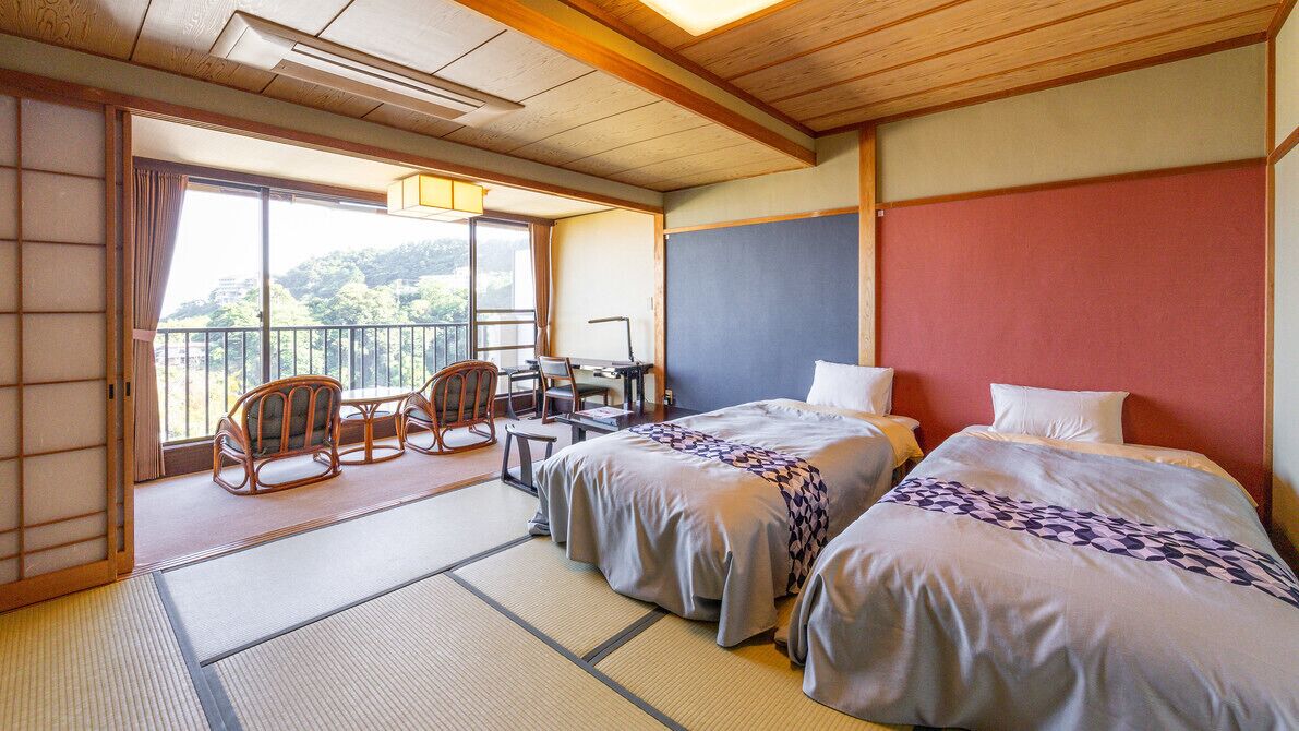 Una habitación japonesa tradicional con suelo de tatami, dos camas con almohadas, un escritorio con sillas y un balcón con vistas a las montañas.