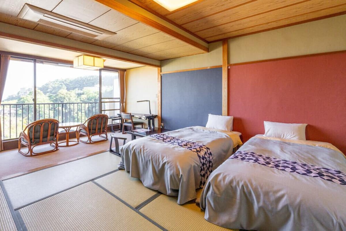 A traditional Japanese room with tatami flooring, two beds with pillows, a desk with chairs, and a balcony with a view of the mountains.