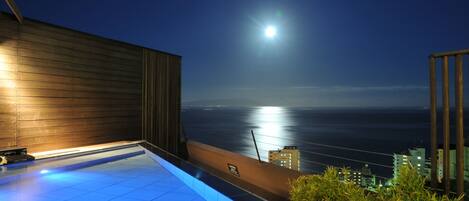 A rooftop pool with a view of the ocean and city skyline at night.