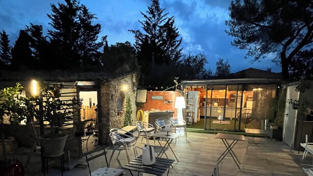 An outdoor patio area with seating and a lamp, surrounded by trees and a stone wall. - Inn4bears (Nîmes)