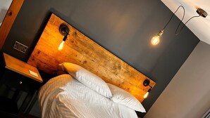 A neatly made bed with white linens and a folded towel, a wooden headboard with a light fixture, a nightstand with a lamp, and a checkered rug on a wooden floor.