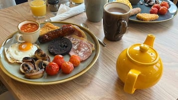 Aamiaispöydällä on monenlaisia ruokia, kuten munia, makkaroita, sieniä ja tomaatteja. Lisäksi pöydällä on kahvikuppi, teekannu ja lasi appelsiinimehua.