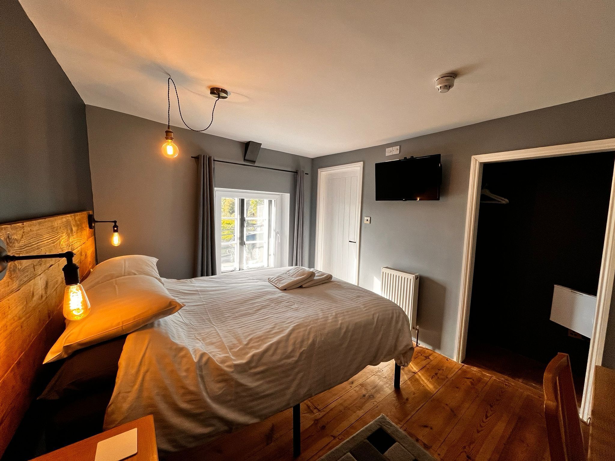 Un dormitorio con cama, lámparas de noche, televisor, radiador y una puerta que da a otra habitación.