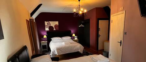 Quarto de hotel com duas camas, uma mesa de cabeceira de madeira com abajur, uma cadeira e uma televisão presa na parede.