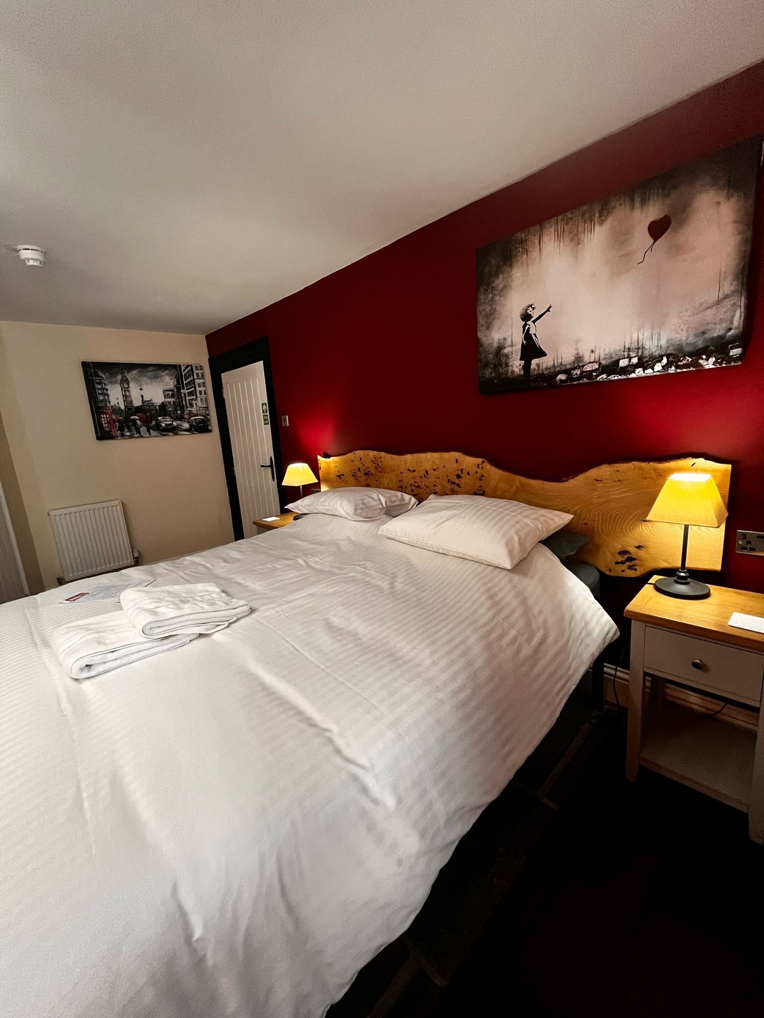 Una cama bien hecha con sábanas blancas, dos lámparas de noche, una mesita de noche de madera y una pared de acento roja con un cuadro.