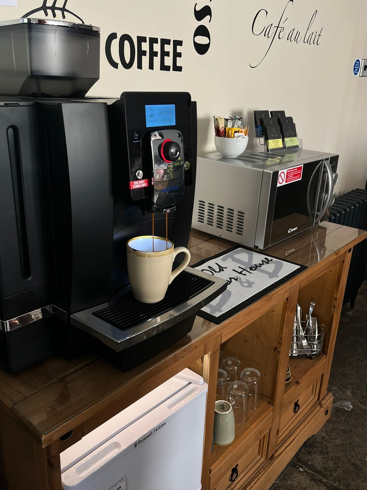 A coffee station with a coffee machine, a cup of coffee, a refrigerator, and a wall sign that says "COFFEE".